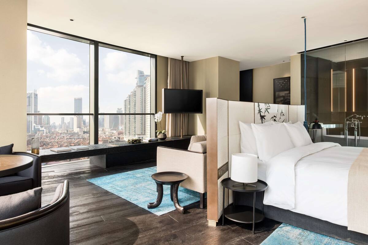 Ein modernes Hotelzimmer mit großem Fenster und Blick auf die Stadt, einem Bett mit weißer Bettwäsche, einem kleinen Beistelltisch und einer Sitzecke mit Stuhl und Tisch.