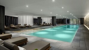 A modern indoor swimming pool area with lounge chairs and a sunbed, surrounded by walls with vertical slats and recessed lighting.