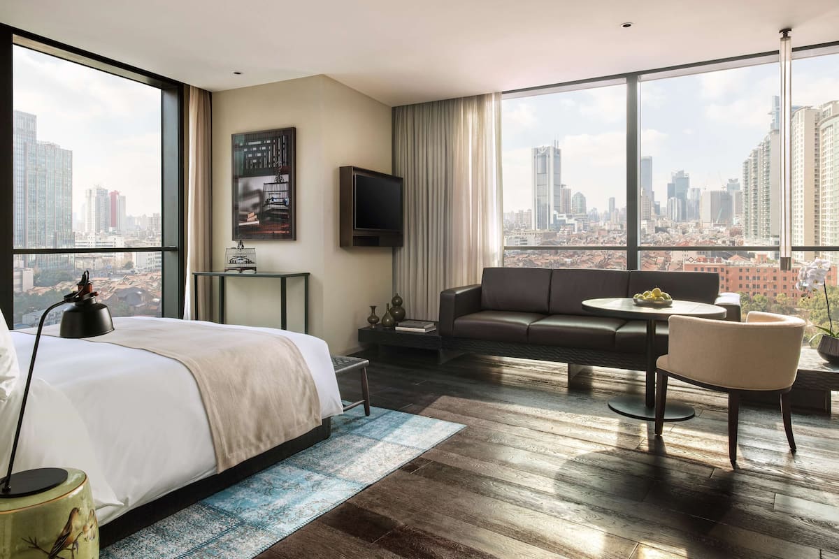 Ein modernes Hotelzimmer mit einem großen Bett, einer Couch, einem Esstisch und einem Fernseher. Das Zimmer verfügt über große Fenster mit Blick auf die Stadt.
