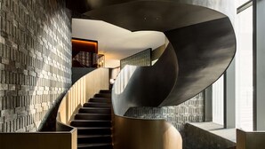 A modern spiral staircase with a brick wall backdrop and a wooden finish.