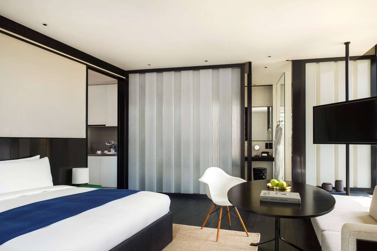 Ein modernes Hotelzimmer mit einem großen Bett, einem Schreibtisch, einem Sessel, einem Fernseher und einem durch eine Schiebetür einsehbarer Badezimmer.