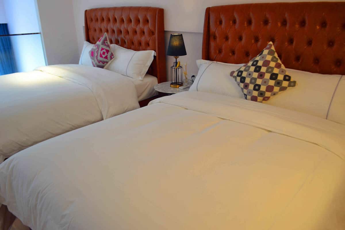 Una habitación de hotel con dos camas, cada una con cabecero capitonado, ropa blanca y almohadas decorativas. Una pequeña mesa con lámpara se encuentra entre las camas.