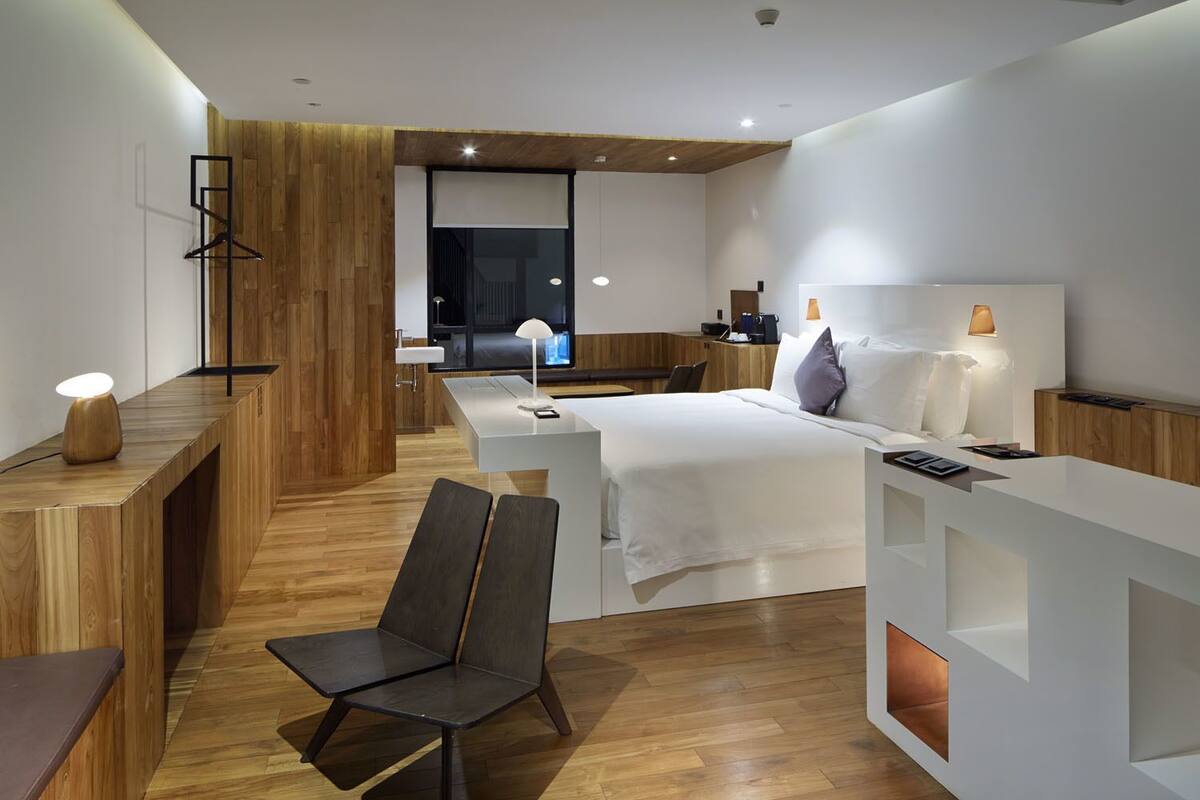 Une chambre d’hôtel moderne, équipée d’un lit, de tables de chevet, d’un bureau, d’une chaise et d’une salle de bain visible à travers une porte en verre.