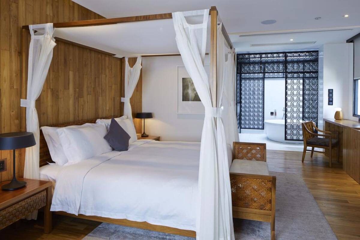Un lit à baldaquin avec des draps blancs, une tête de lit en bois et un ciel de lit. On aperçoit une table de chevet avec une lampe, une chaise et une petite table avec une autre chaise. Une grande fenêtre offre une vue sur l’extérieur.
