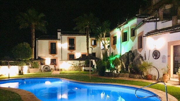Une piscine entourée d’une terrasse bien éclairée, agrémentée de palmiers et bordée d’un bâtiment dont le toit est recouvert de tuiles.