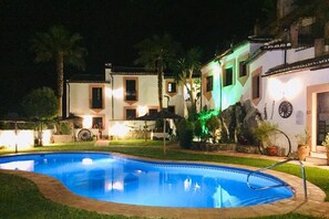 A swimming pool surrounded by a well-lit patio area with palm trees and a building with a tiled roof. - Casas de Montejaque (Montejaque)