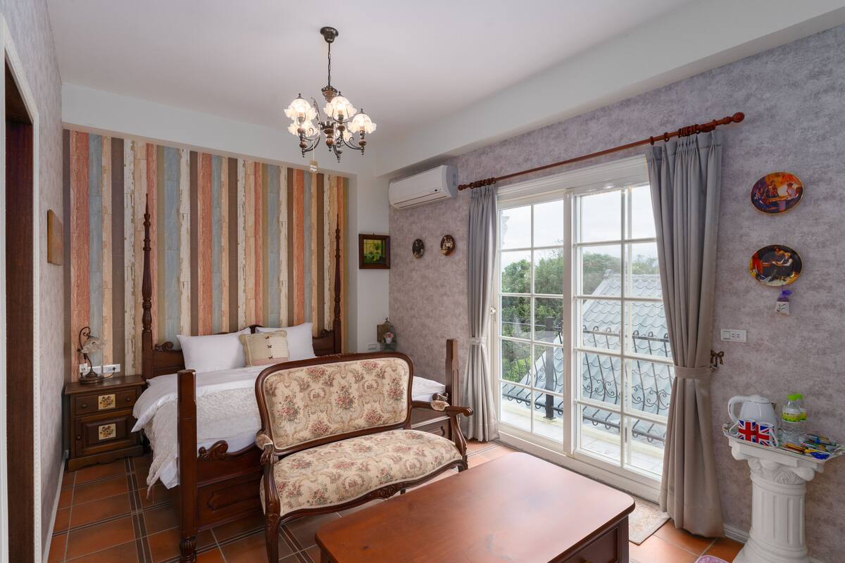 Un dormitorio con cama, sofá, mesa de madera, candelabro y ventana con cortinas.