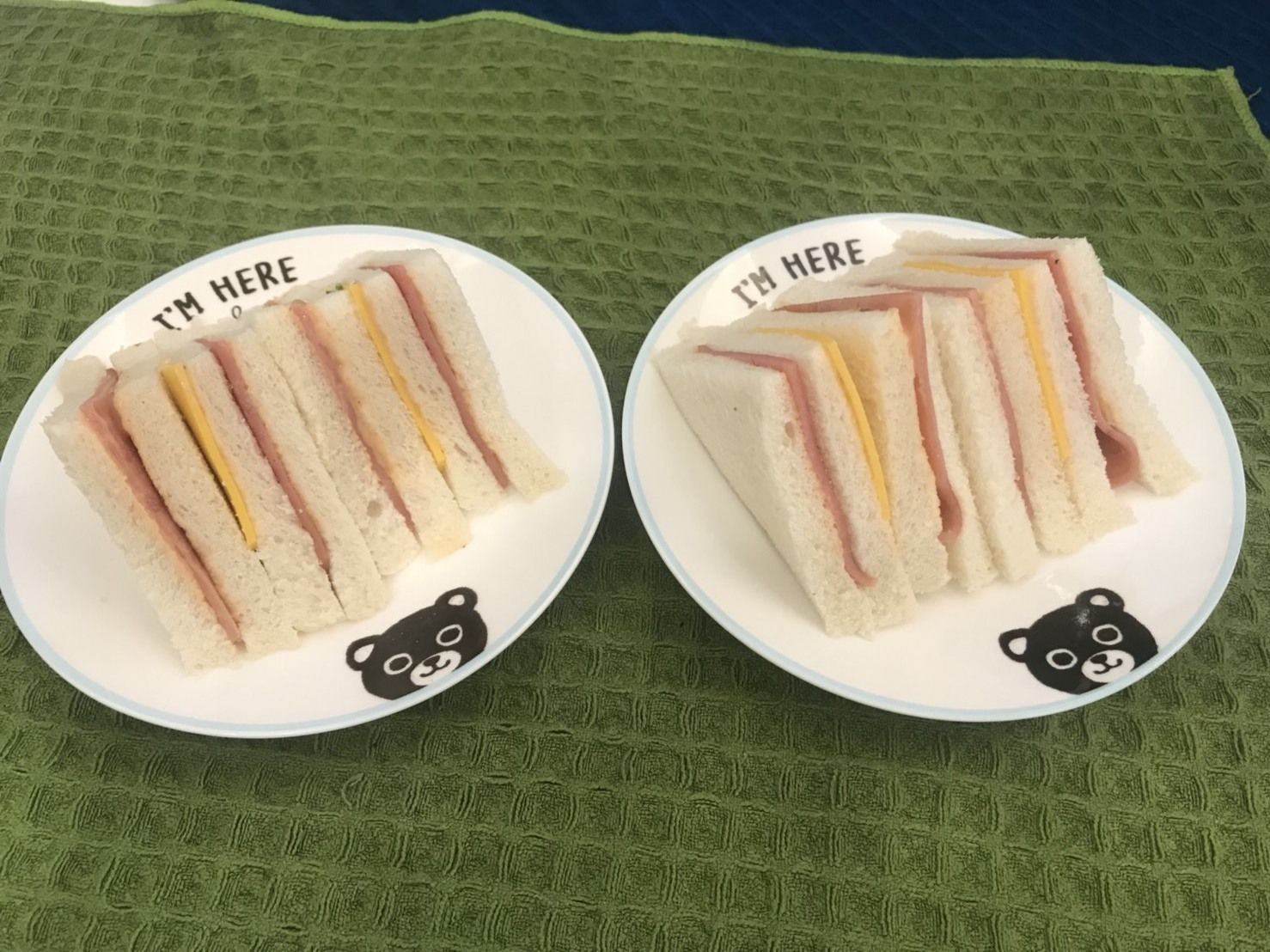 Two plates of sandwiches with 'I'M HERE' text and bear faces, on a green textured surface.