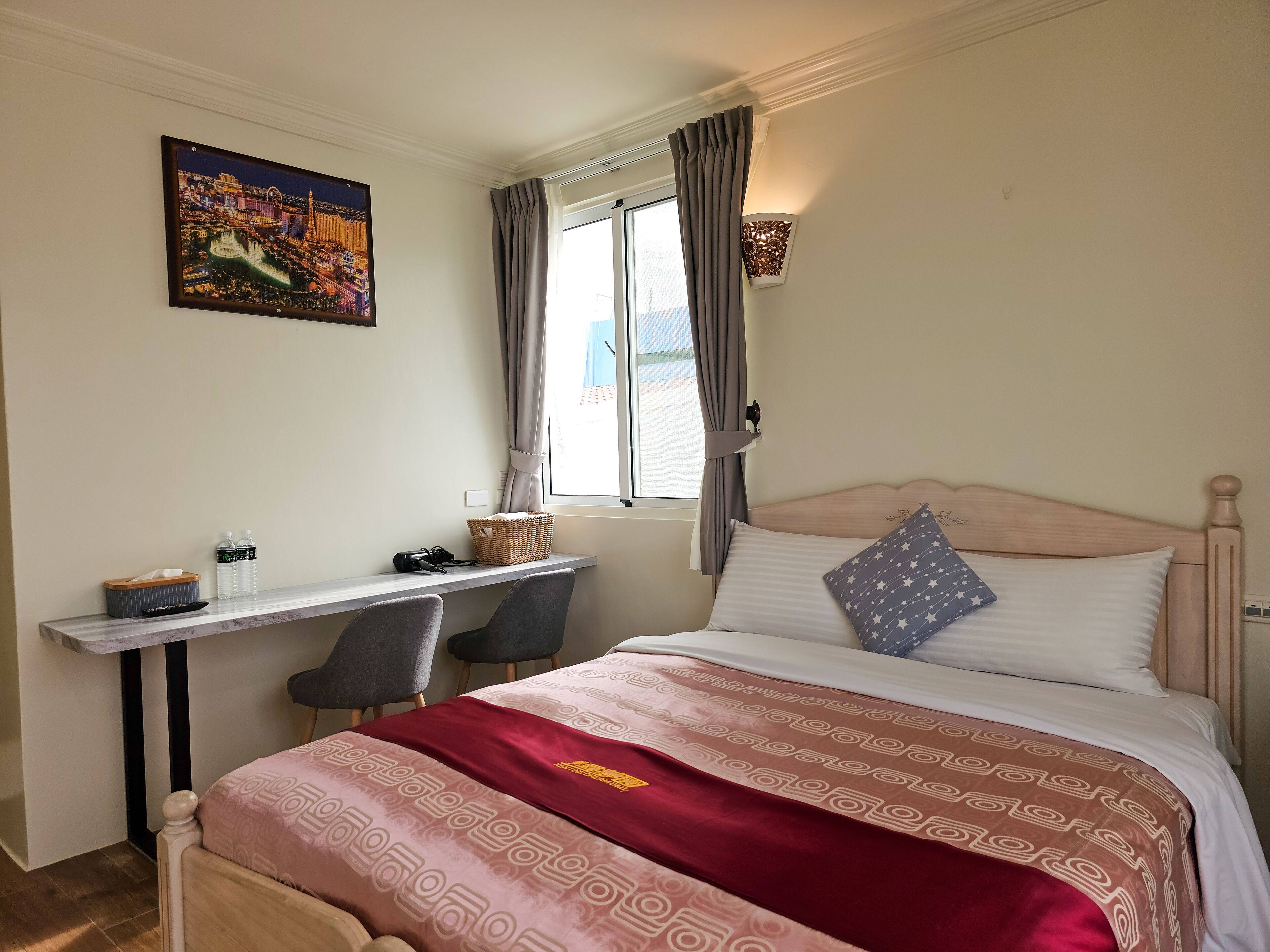A hotel room with a bed, a desk with a chair, a window with curtains, and a wall-mounted lamp.