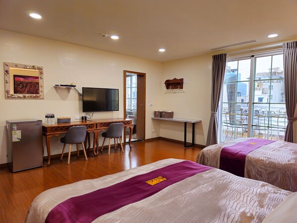 Habitación de hotel con dos camas, un escritorio con sillas, televisor, refrigerador y ventana con vista a edificios.
