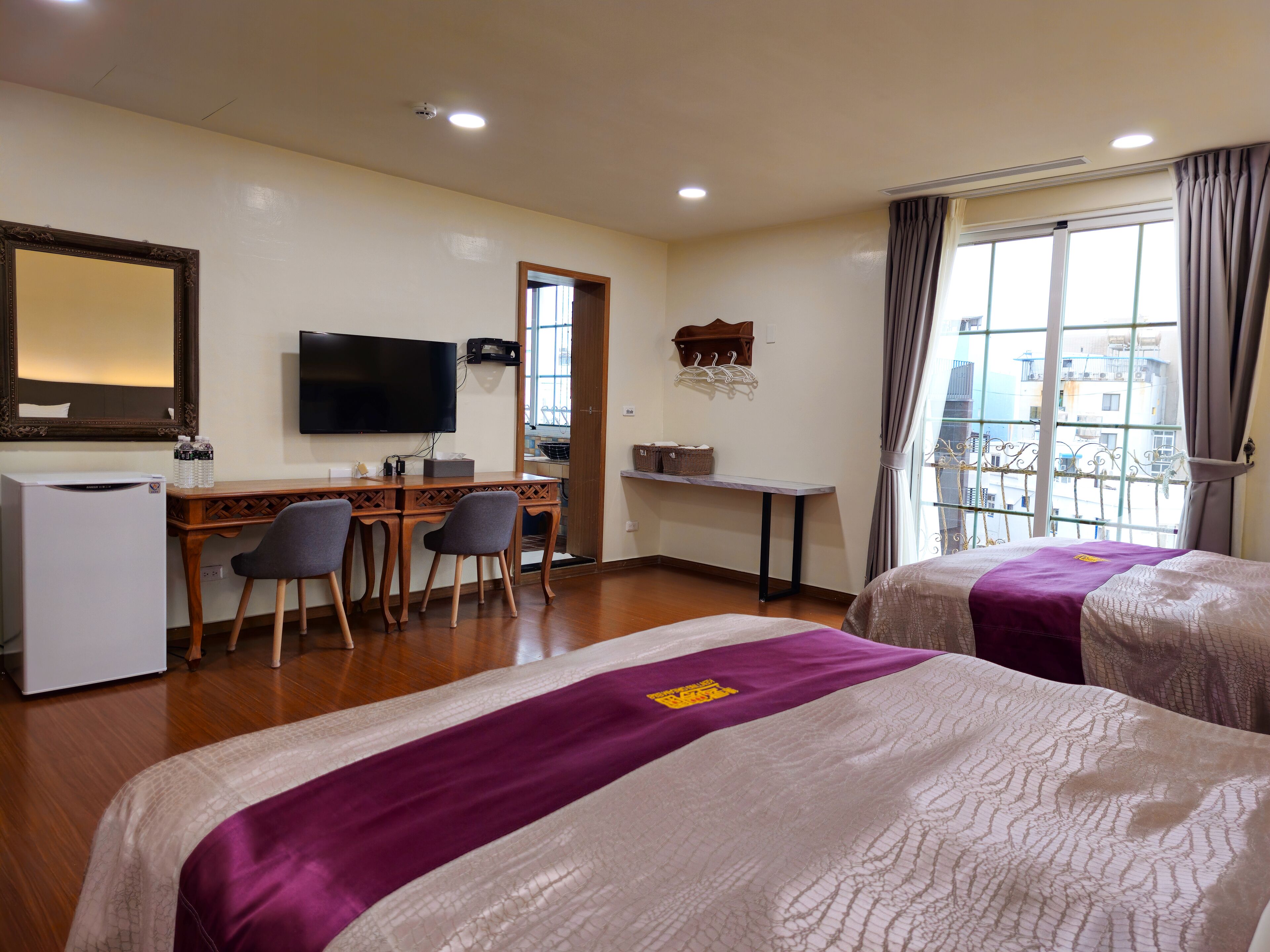 Habitación de hotel con dos camas, un escritorio con silla, televisor, refrigerador y ventana con cortinas.