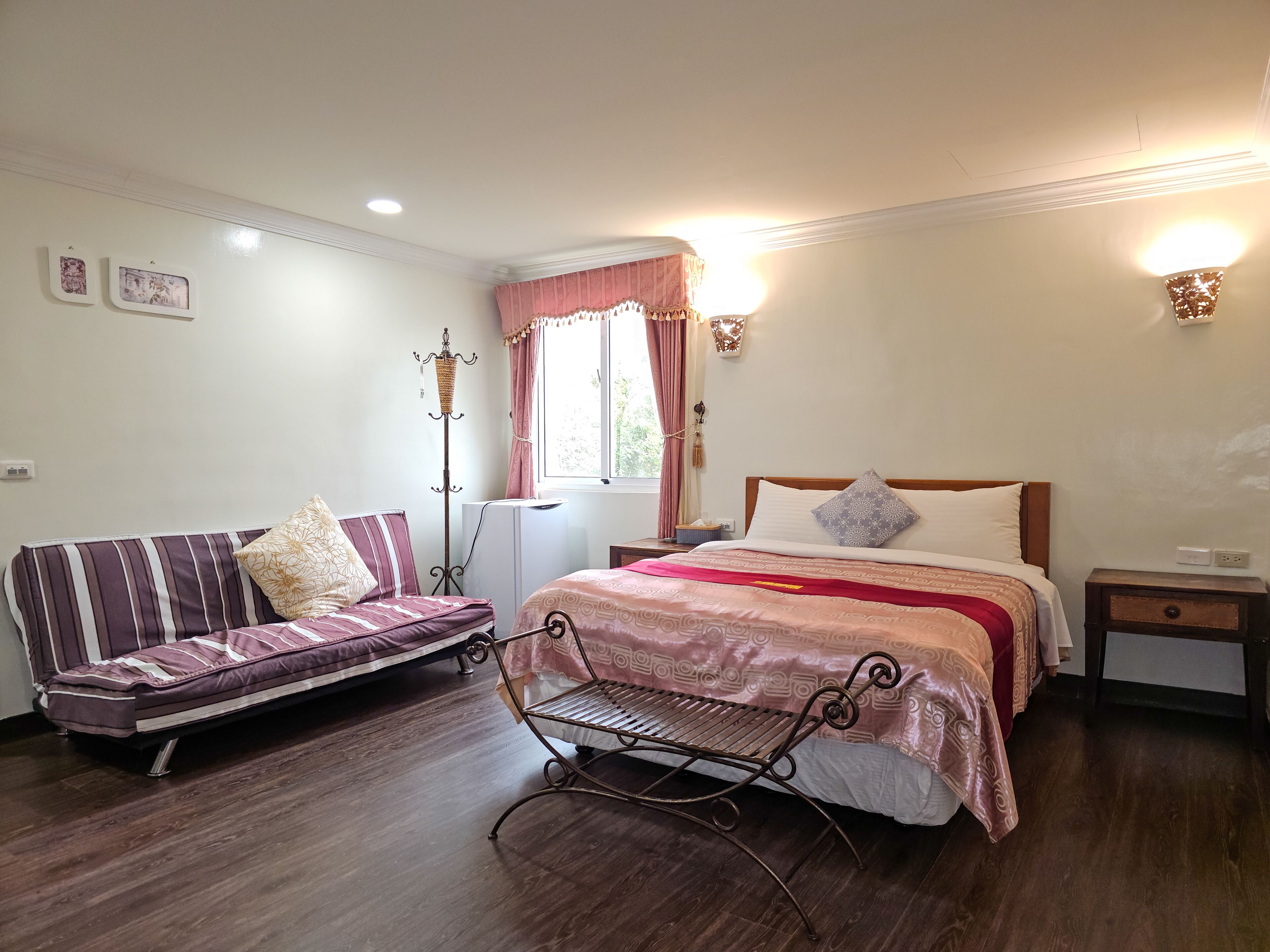 A bedroom with a bed, a sofa, a chair, a standing heater, and a window with curtains.
