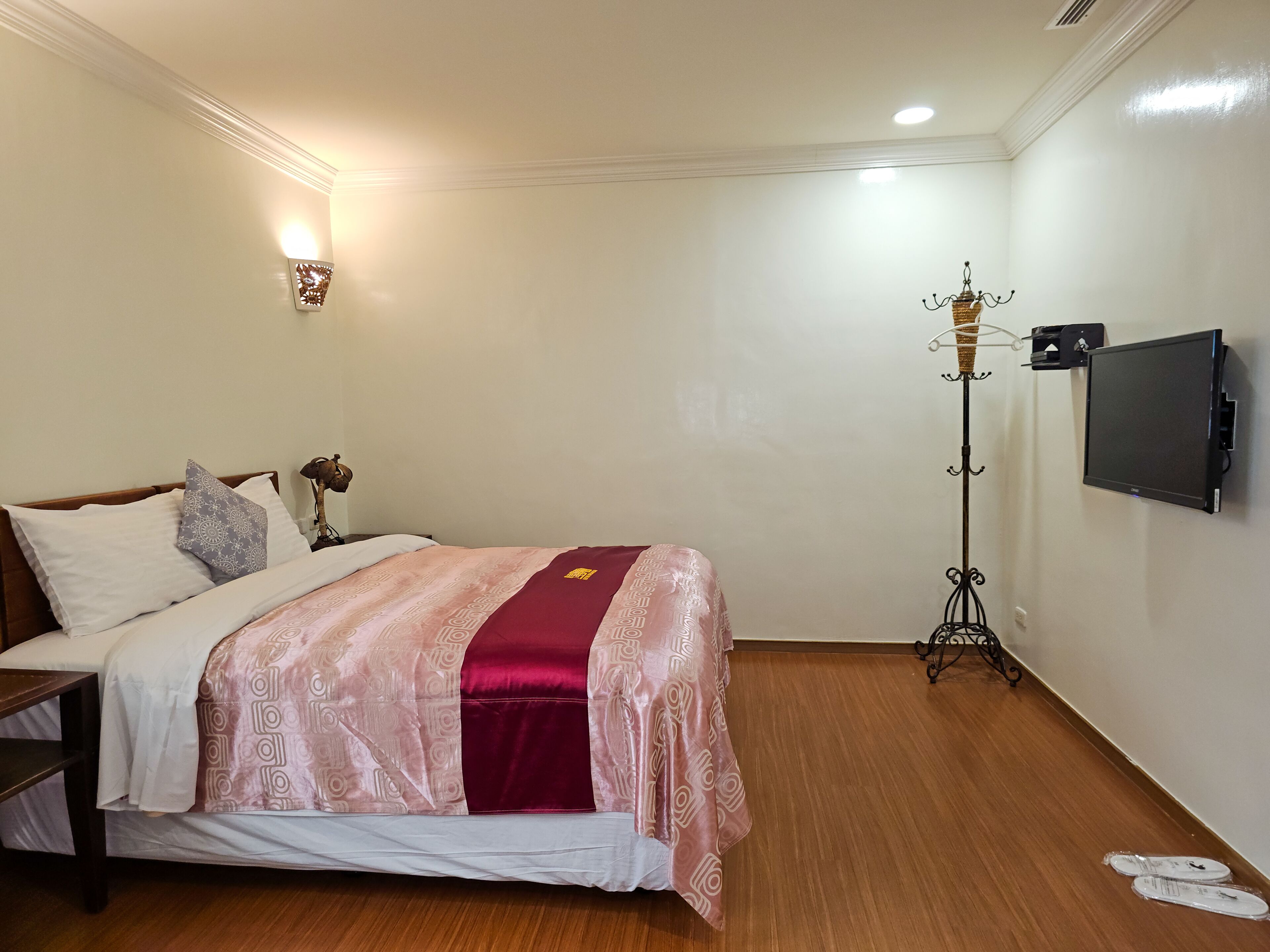 Cama bem arrumada com colcha estampada e almofadas, mesa de cabeceira com abajur, luminária de chão e televisão na parede.