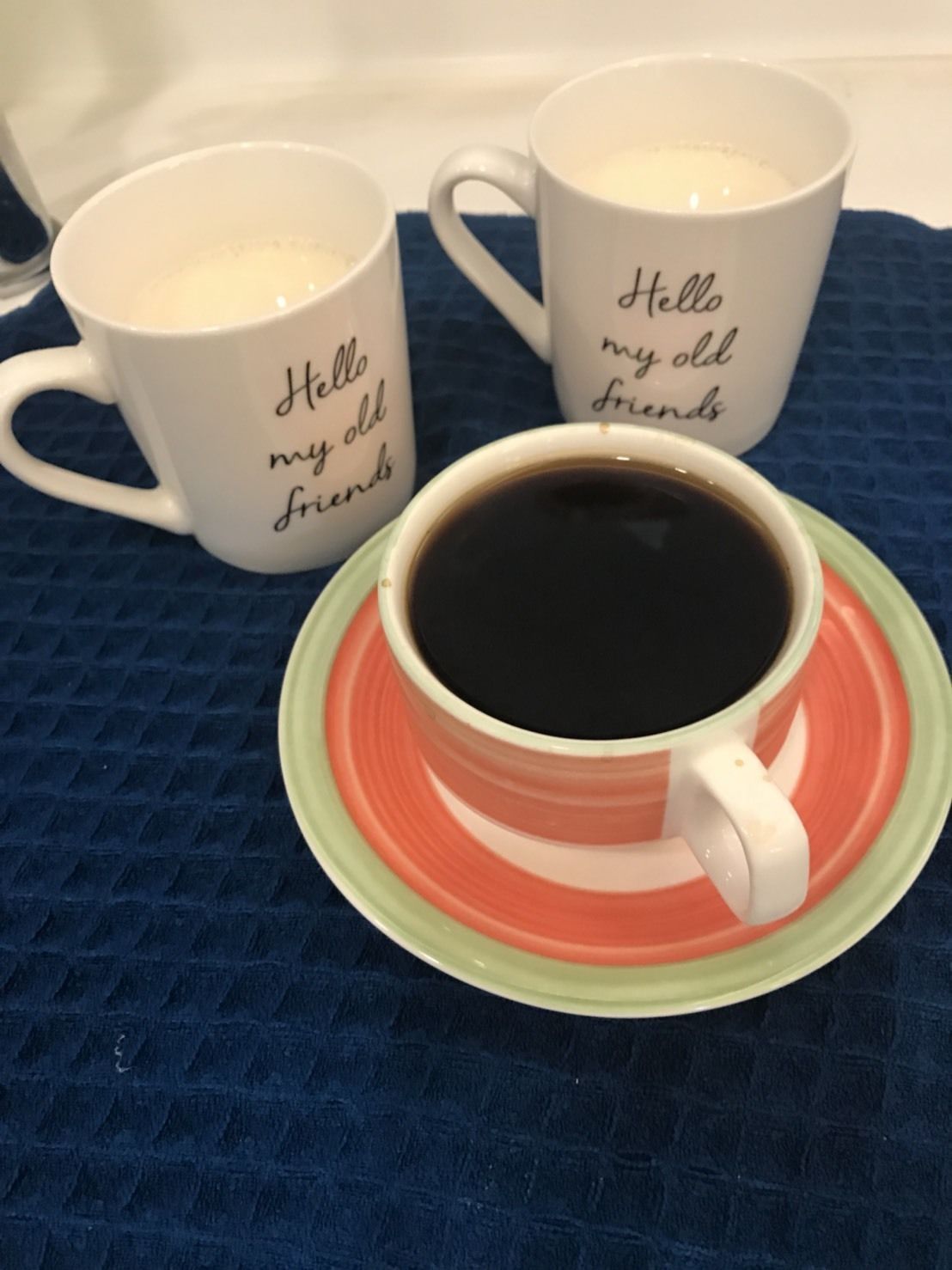 Dos tazas blancas con el texto "Hello my old friends", una taza de café negro sobre un platillo verde y rojo, y un mantel azul con textura.