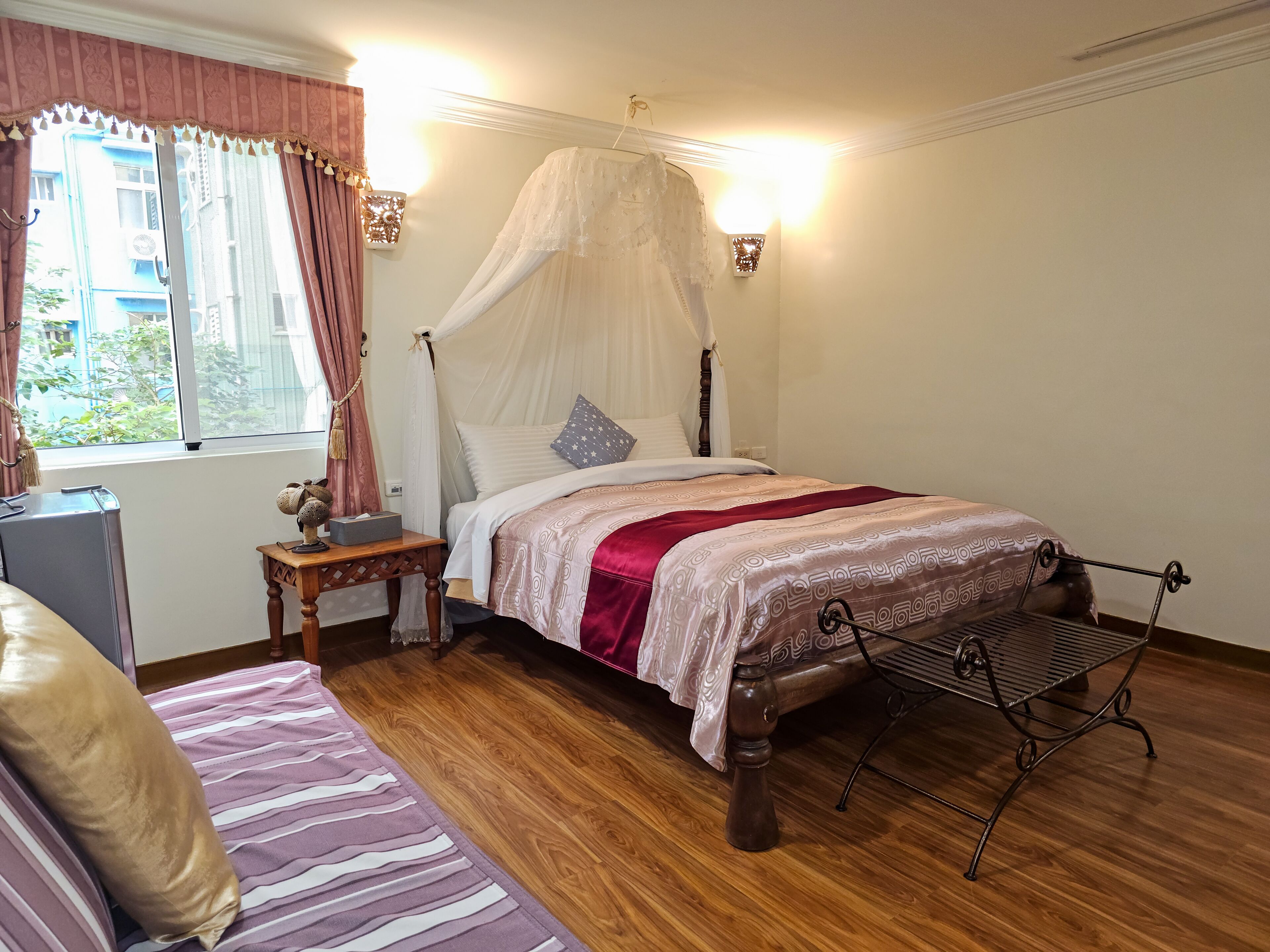 A bedroom with a four-poster bed, a bedside table, a nightstand, a bench, and a window with curtains.