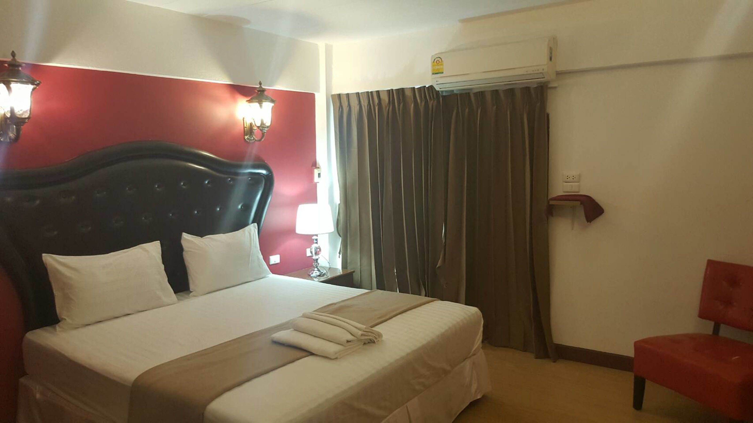 Habitación de hotel con una cama grande, una pared roja, una ventana con cortinas, un equipo de aire acondicionado y una silla roja.