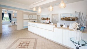 A reception area with a wooden reception desk, a clock, and a receptionist. There are hanging pendant lights, a glass table, and a decorative vase with branches. - Le Dune Beach Club (Messina)