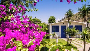 A building with a blue door and windows, surrounded by a garden with pink flowers and palm trees. - Le Dune Beach Club (Messina)