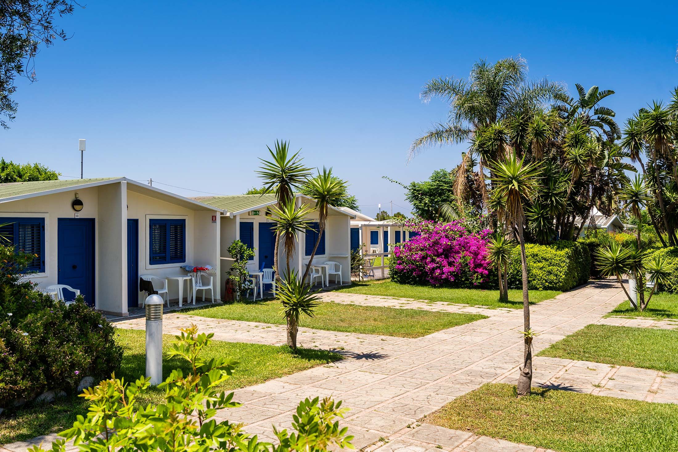 Un hotel con diverse porte blu, posti a sedere all'aperto e un giardino curato con palme e arbusti.