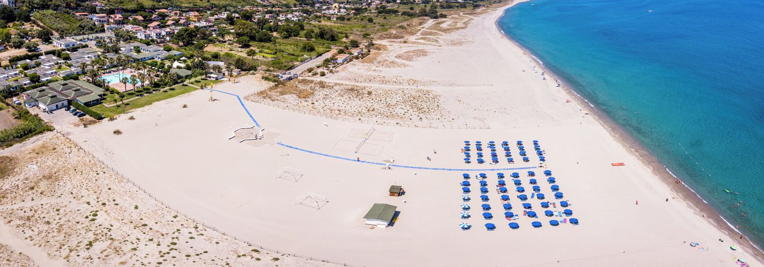Una vista aerea di una spiaggia con una fila di ombrelloni blu, una zona sabbiosa con una recinzione e un'area residenziale sullo sfondo.