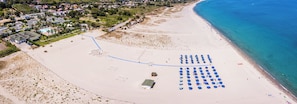 An aerial view of a beach with a row of blue umbrellas, a sandy area with a fence, and a residential area in the background. - Le Dune Beach Club (Messina)