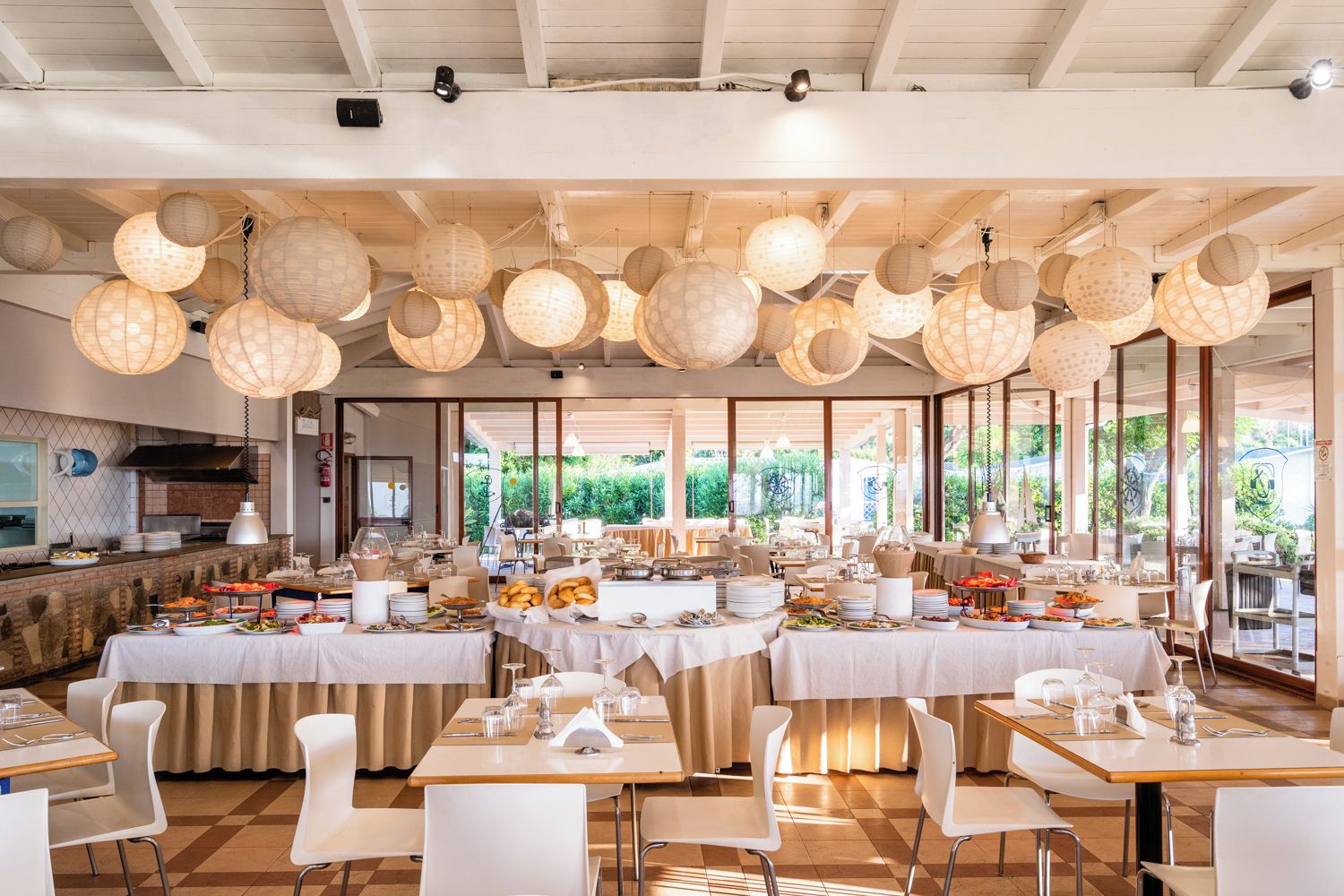 Ampia sala da pranzo con diversi tavoli apparecchiati per un pasto, circondata da ampie finestre che si affacciano su un giardino.