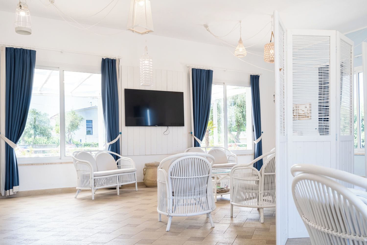 A room with white wicker furniture, a flat-screen TV, and large windows with blue curtains.