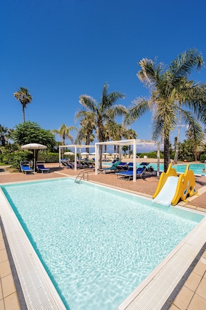 A clear blue pool with lounge chairs and umbrellas, surrounded by palm trees.