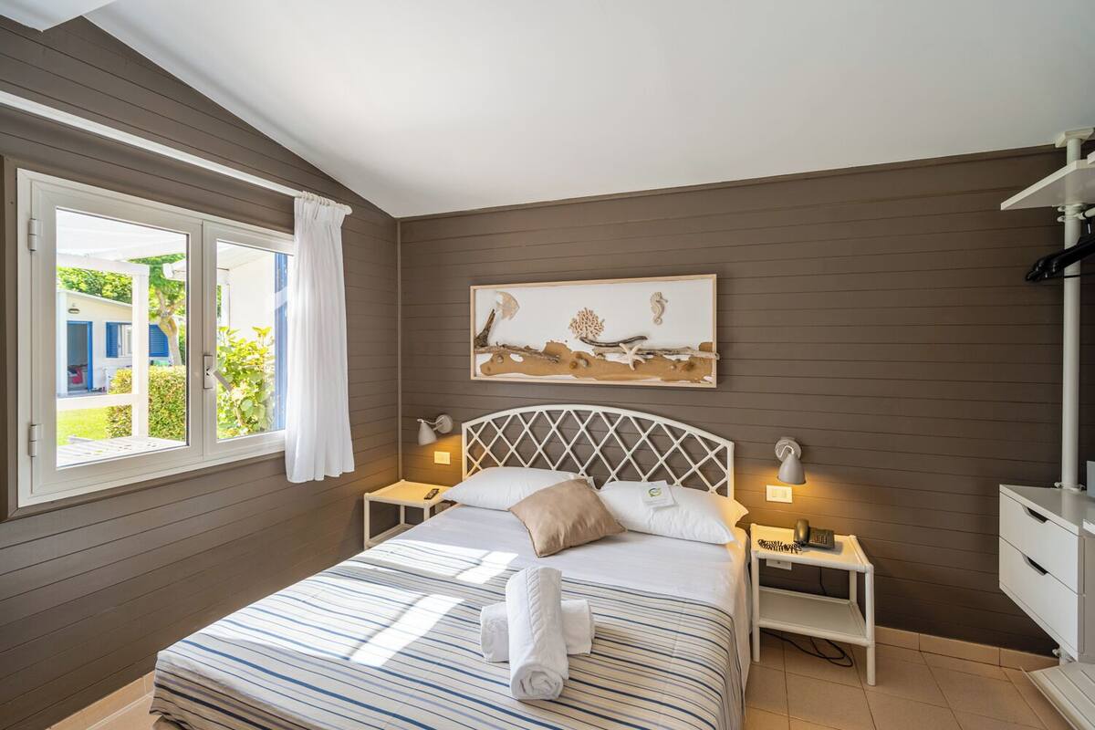 A bedroom with a bed, bedside tables, a nightstand, a window with curtains, and a wall-mounted artwork.