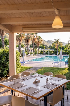 An outdoor dining area with tables set for a meal, overlooking a swimming pool and palm trees. - Le Dune Beach Club (Messina)