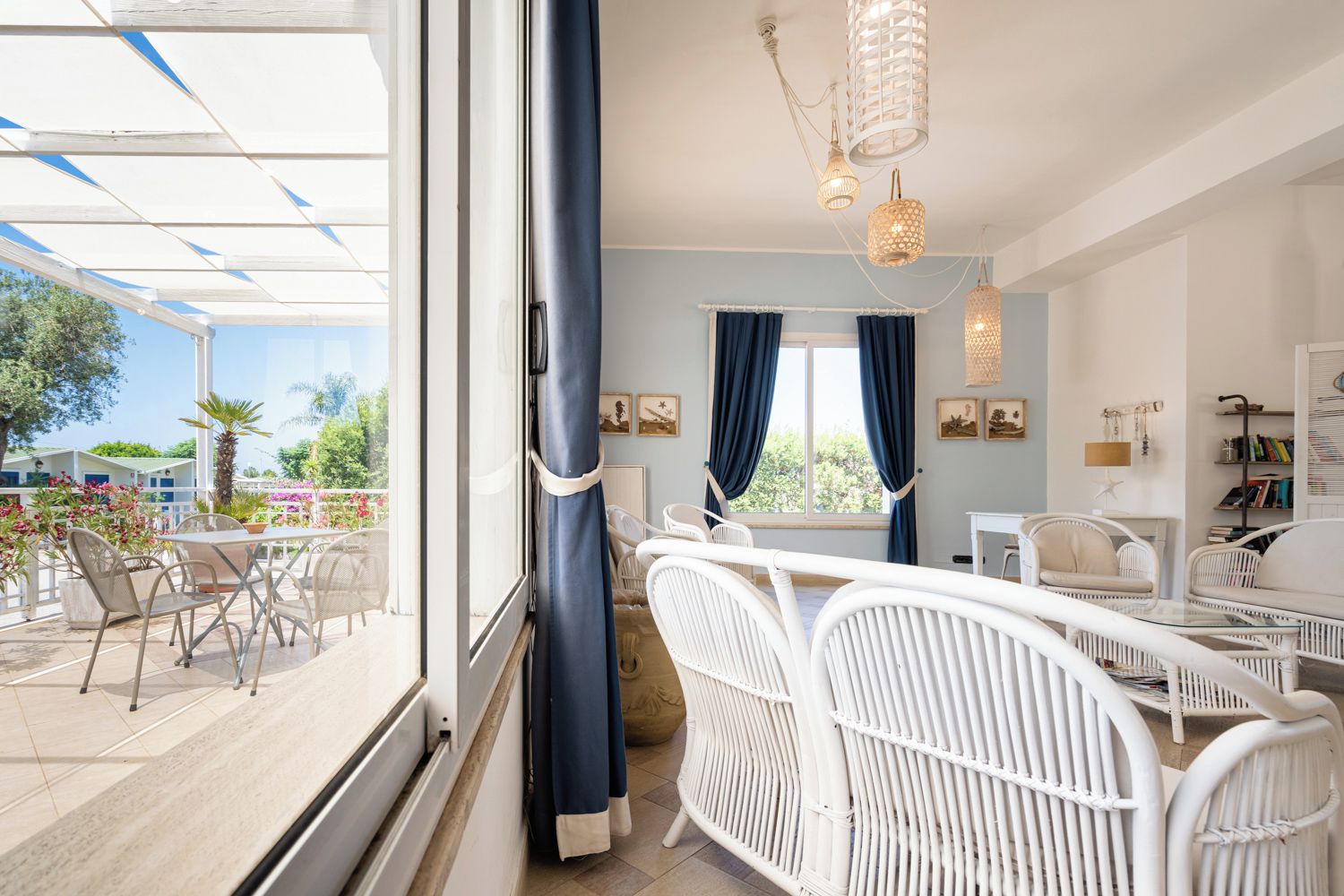Una stanza con balcone, mobili in vimini bianco, un'ampia finestra con tende e un lampadario.