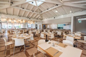Un vaste espace dédié à la salle à manger, avec de nombreuses tables et chaises, un plafond haut avec des poutres apparentes et une cuisine ouverte.