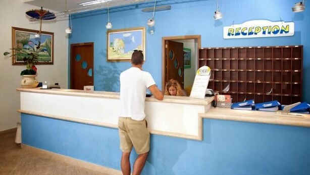 A reception desk with a blue wall, a 'RECEPTION' sign, and a man standing behind it. - Le Dune Beach Club (Messina)