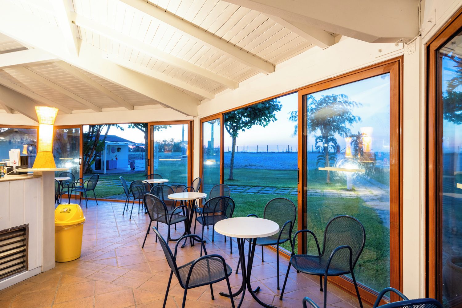 Un'area patio interna con tavoli e sedie, ampie porte vetrate che danno su un corpo d'acqua e un cestino giallo.