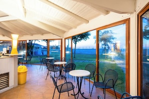 An indoor patio area with tables and chairs, large glass doors leading to a view of a body of water, and a yellow trash bin. - Le Dune Beach Club (Messina)