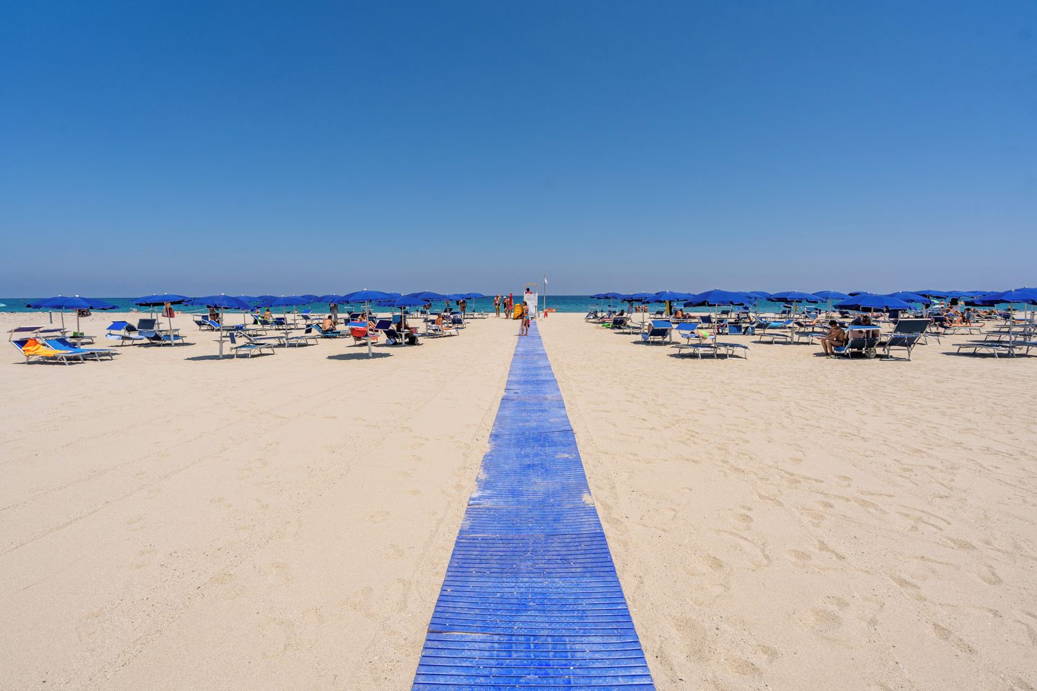 Una spiaggia con una passerella blu che conduce all'acqua, circondata da lettini e ombrelloni.