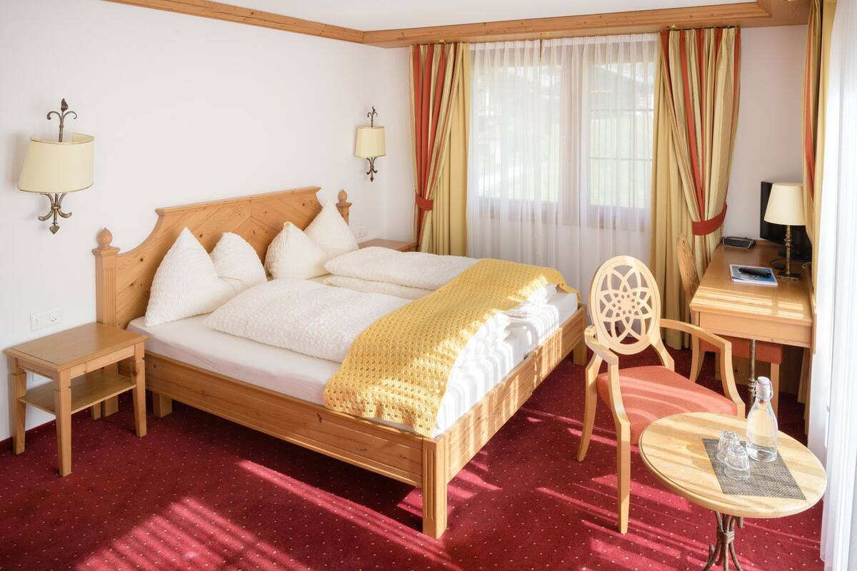 A hotel room with a wooden bed, two bedside tables, a desk with a chair, and a small round table with glasses.