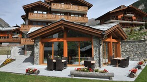 A multi-story wooden chalet with balconies, a glass-enclosed patio area, and stone walls. - Hotel Mountain Paradise (Zermatt)