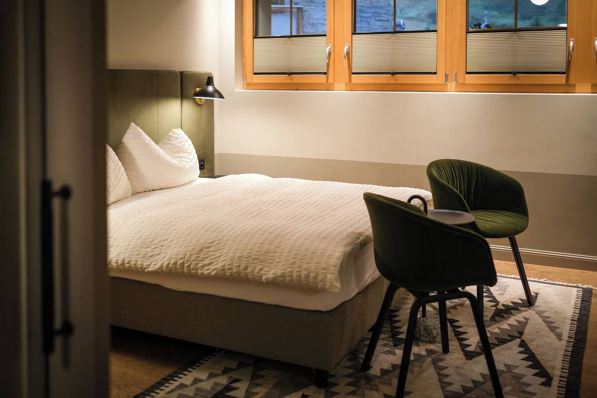 A bedroom with a bed, two chairs, a window with blinds, and a patterned rug.