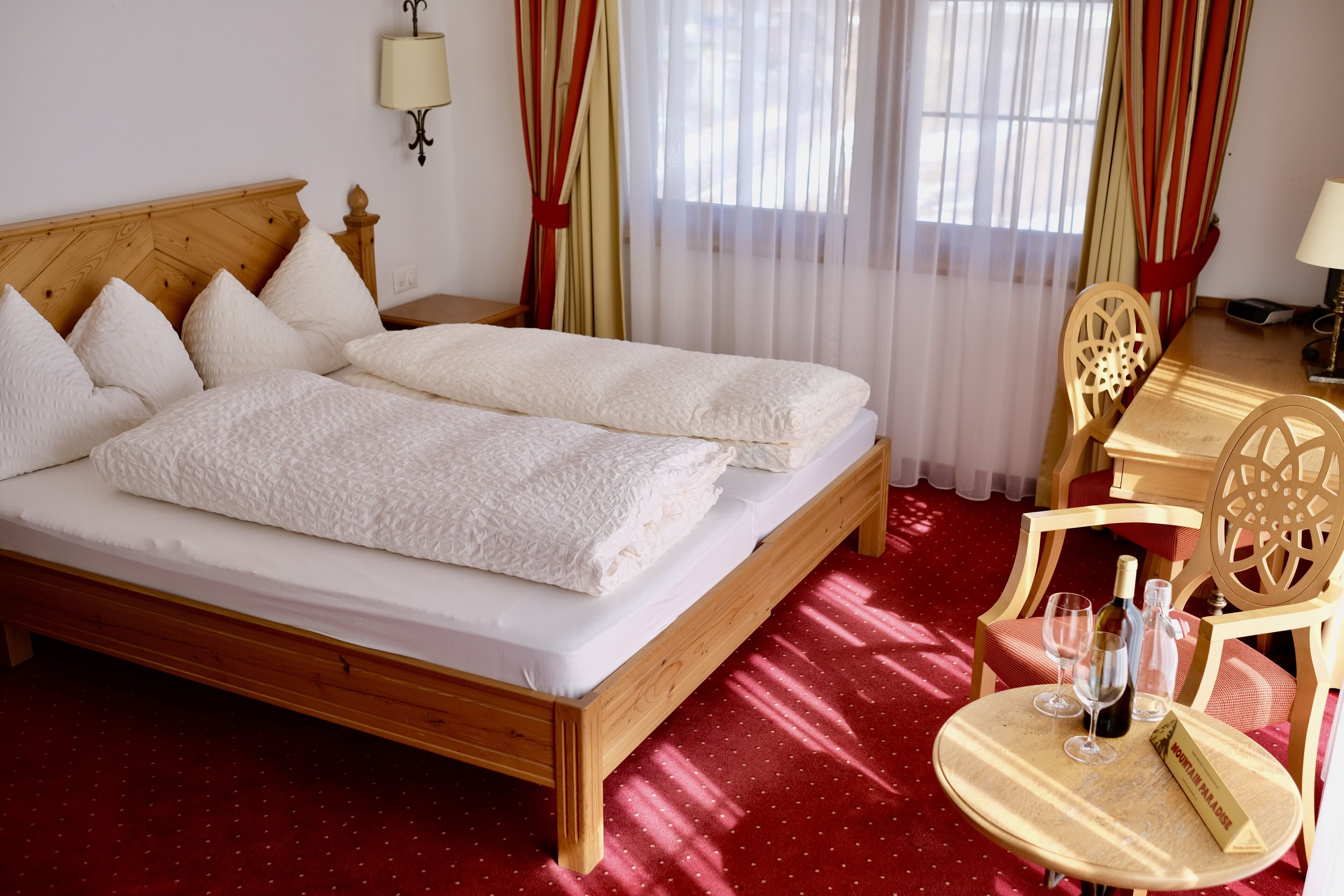 A neatly made bed with white linens, a wooden headboard, and a red carpeted floor. There is a small round table with a bottle and glasses, and a chair with a wooden design.