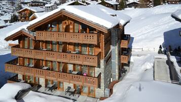 A multi-story wooden chalet with balconies, a ski slope in the background, and snow-covered surroundings.