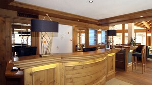 A wooden reception desk with a curved design, a large mirror, and a wall-mounted lamp. - Hotel Mountain Paradise (Zermatt)