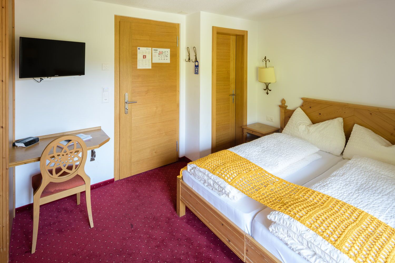 A hotel room with a bed, a desk, a chair, a TV mounted on the wall, and a door with a notice.