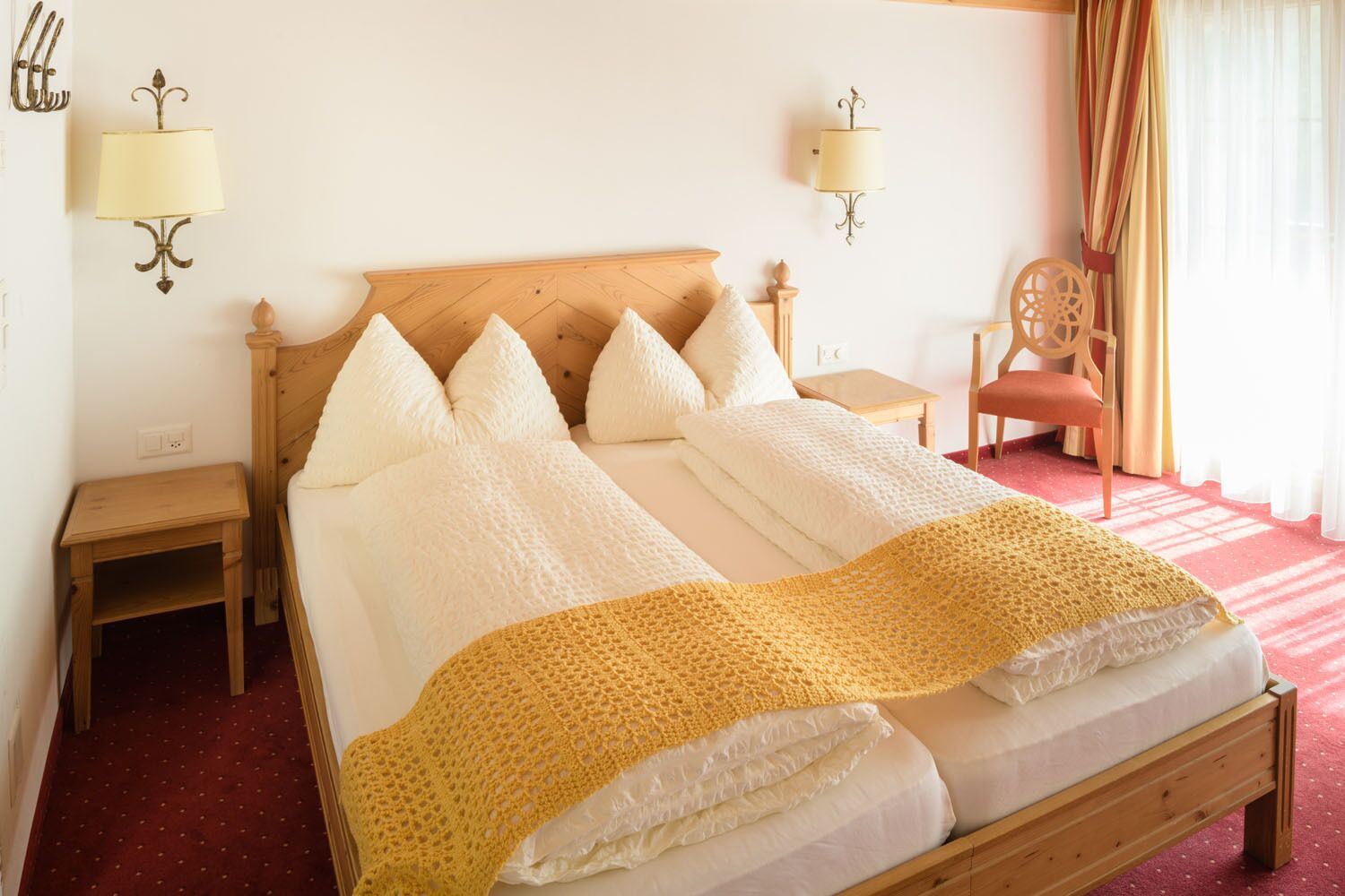 Habitación de hotel con cama de madera, ropa de cama blanca, manta amarilla, dos mesitas de noche, una silla y lámparas fijadas a la pared.