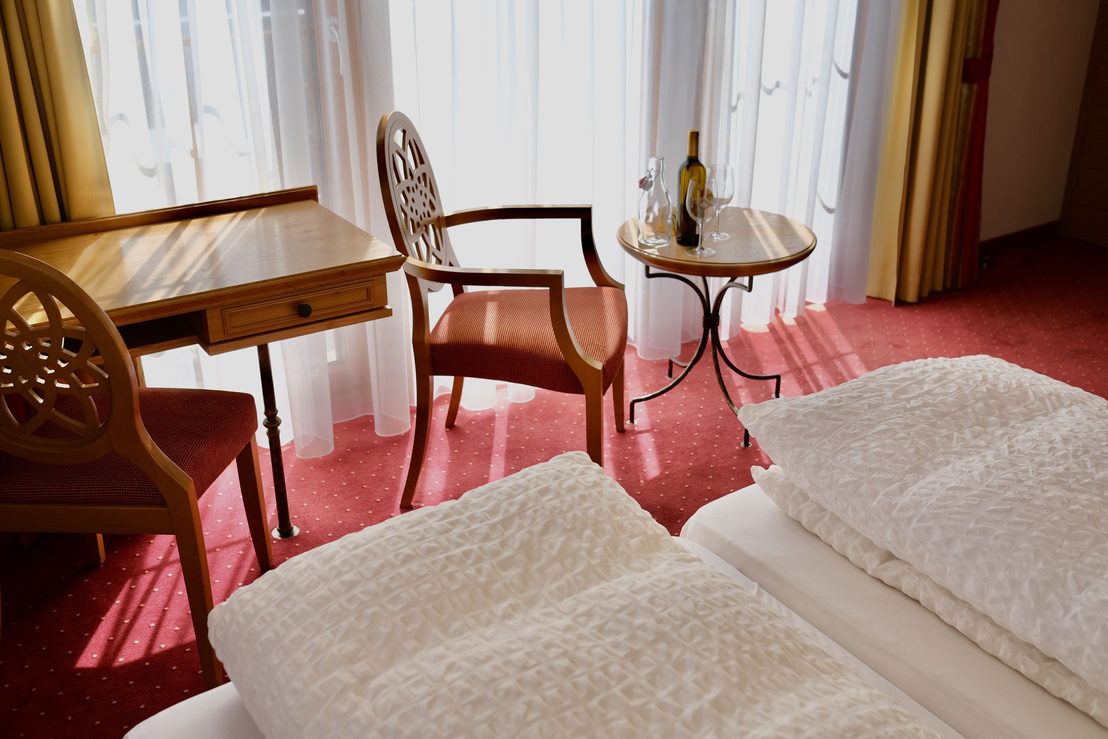 Quarto de hotel com uma escrivaninha de madeira, uma cadeira, uma mesa redonda pequena com uma garrafa e copos, e duas camas com roupa de cama branca.