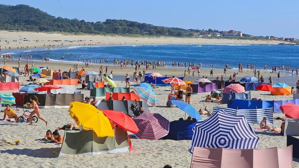 Una playa con tiendas de campaña y sombrillas, gente tomando el sol y nadando, con vistas despejadas al mar y colinas distantes.