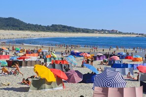 Une plage avec des tentes et des parasols, des gens qui prennent des bains de soleil et se baignent, et une vue dégagée sur la mer et les collines lointaines.