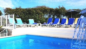 Una piscina con sillas de descanso azules y blancas, una fuente de agua y un cielo despejado.
