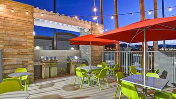 Une terrasse extérieure avec un espace barbecue, des chaises vertes et des parasols rouges.
