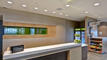 Zona de recepció d'hotel amb un disseny modern, que inclou un taulell de recepció, una pantalla digital i una màquina expenedora.